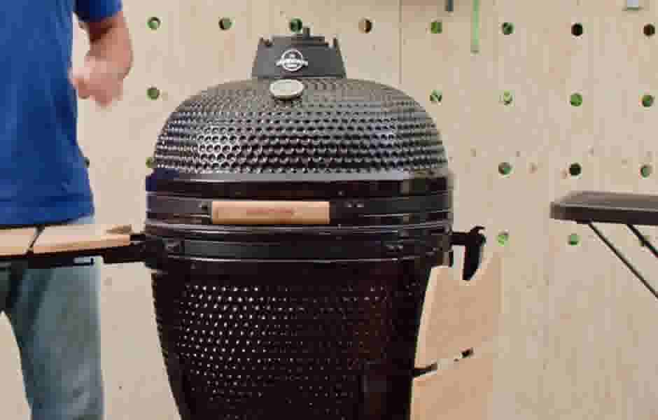Geopende zwarte keramische kamado barbecue met vis en vlees op het rooster, omringd door houten snijplanken en keukengerei in een tuin.
