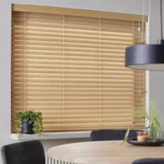 Houten jaloezie in lichtbruine kleur voor een raam, met een ronde houten tafel en groene plant op de voorgrond.