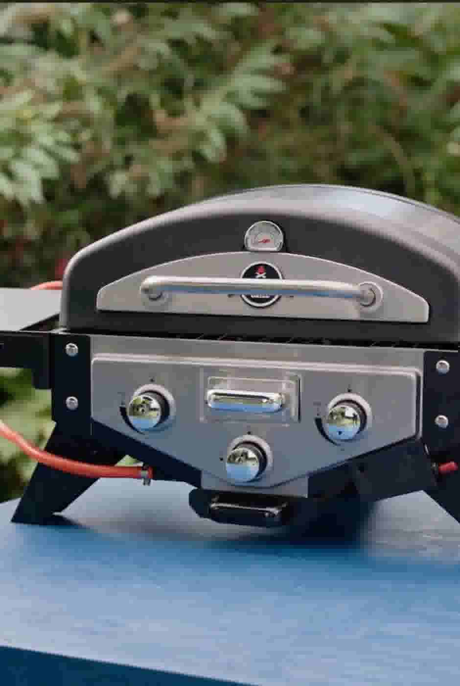 Compacte gasbarbecue met temperatuurmeter, bedieningsknoppen en metalen handgreep, geplaatst op een blauwe tafel in de tuin.
