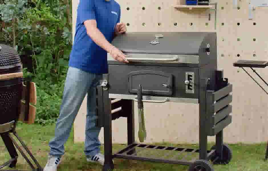 Persoon in blauw shirt opent een zwarte barbecue in een tuin met houten schutting en groene planten op de achtergrond.