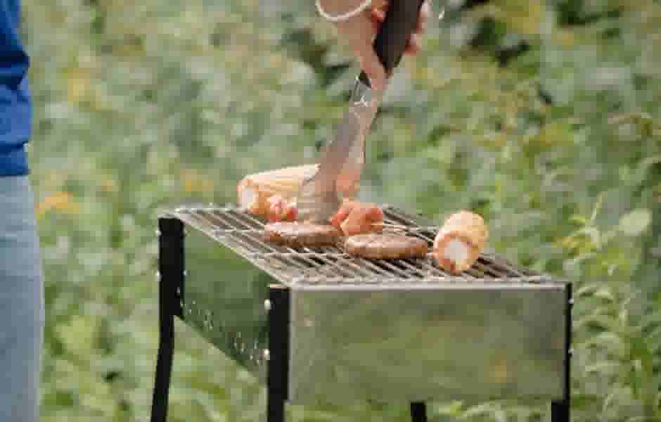 Persoon draait vlees en maïskolven om op een open grill barbecue in een groene tuin met een grilltang in de hand.