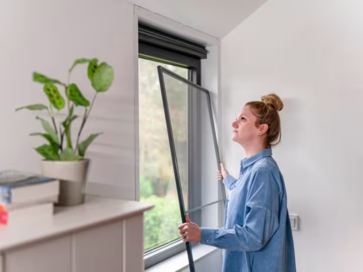 Vrouw installeert een raamhor in een lichte kamer met een groene plant op de vensterbank.