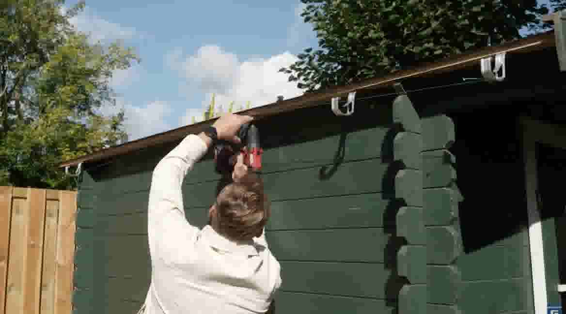 GAMMA klusser gebruikt een rode boormachine om iets te bevestigen aan de dakrand van een groene tuinschuur.