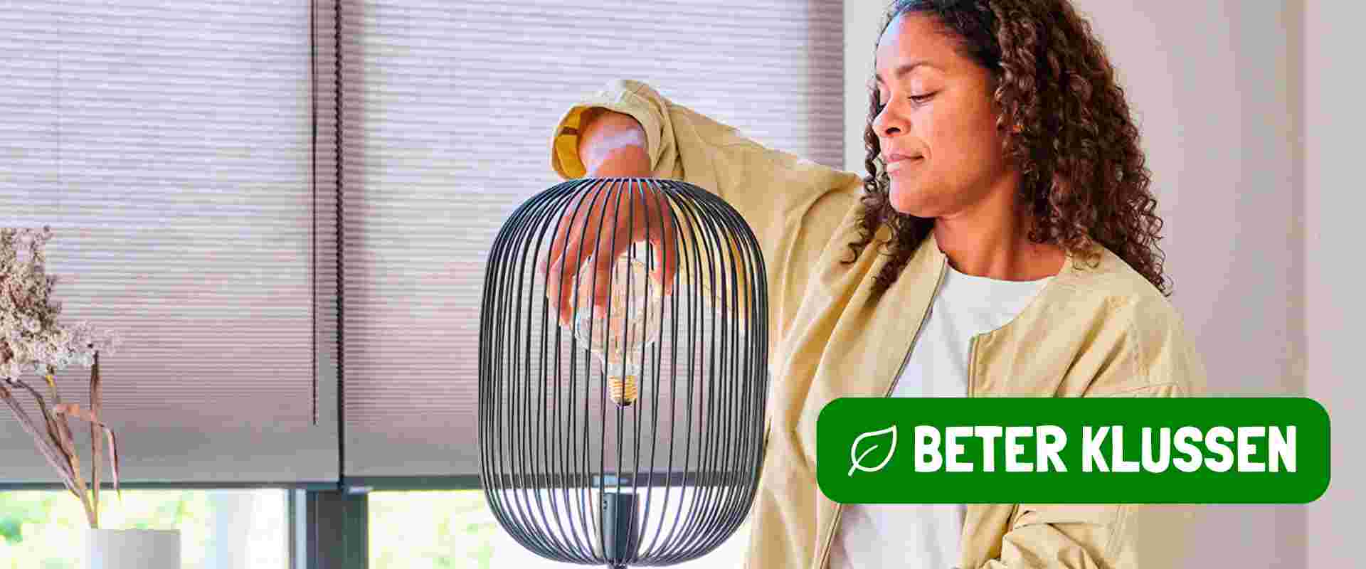 Vrouw in beige jas staat bij een raam met jaloezieën. Ze houdt een zwarte vloerlamp vast en draait er een LED lamp in. 