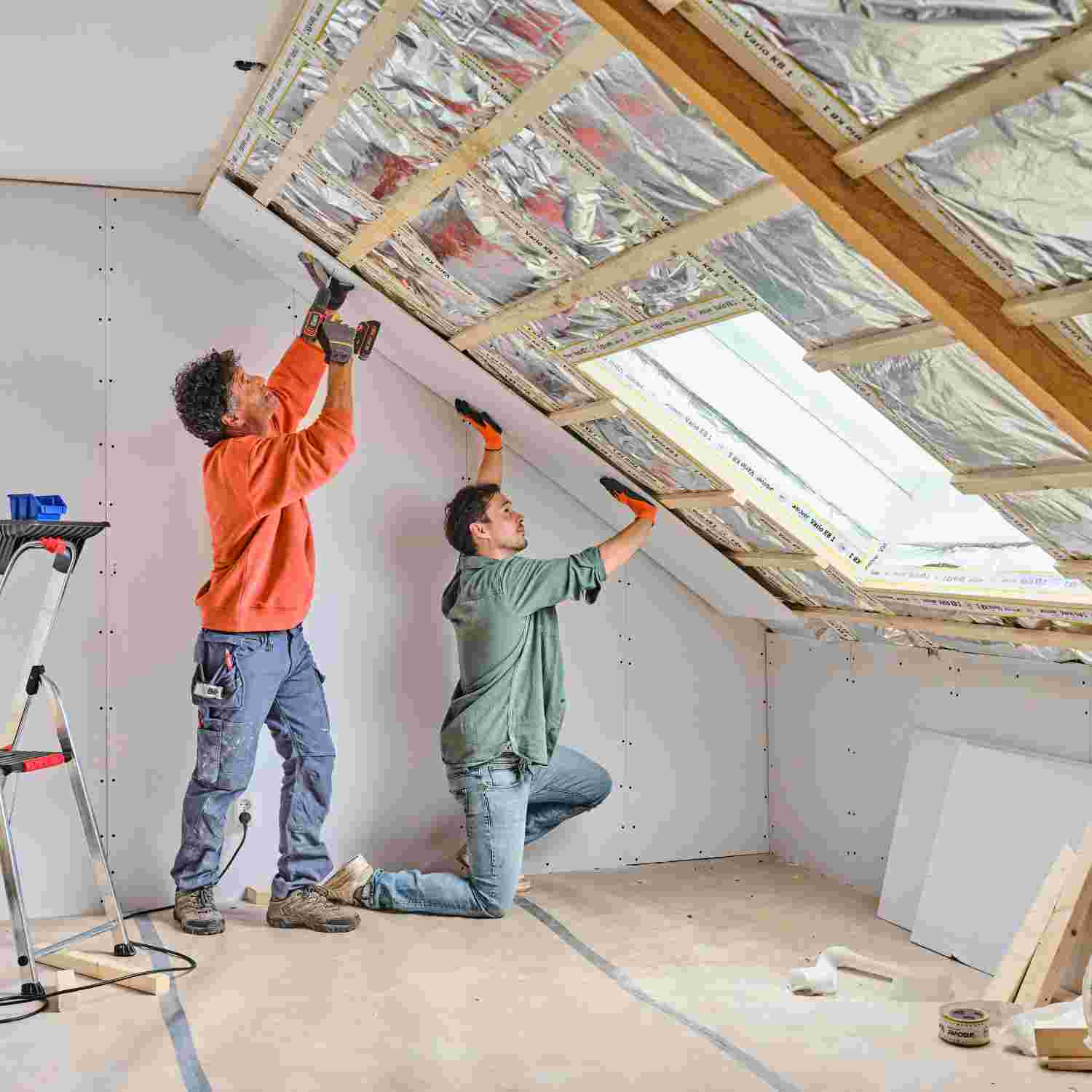 Twee mannen installeren isolatiemateriaal en gipsplaten aan een schuine zolderwand met dakraam.