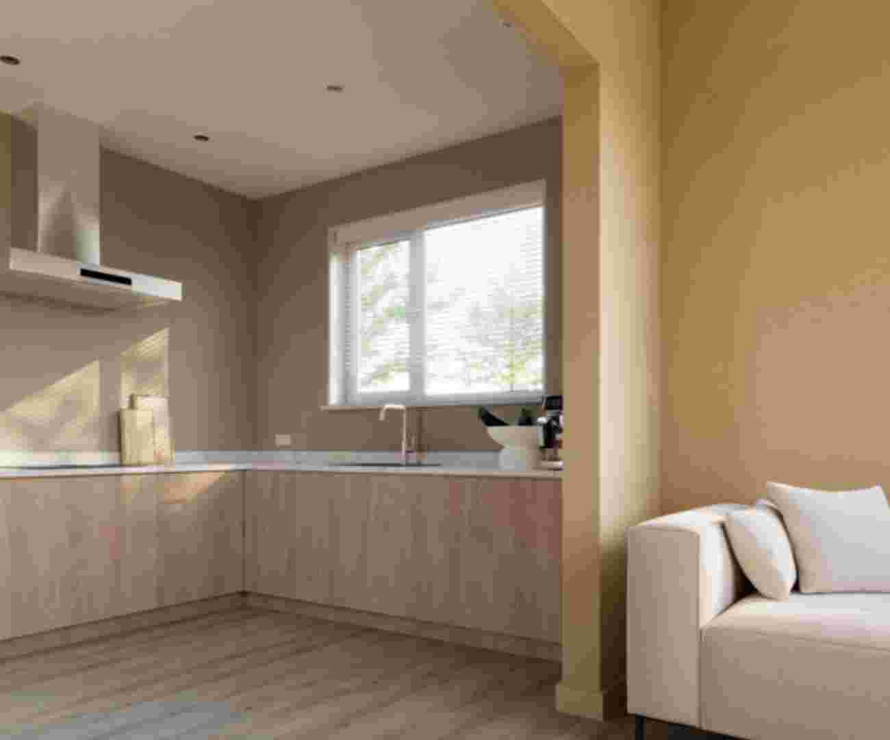 Moderne open keuken met houten kasten, wit werkblad en afzuigkap, naast een lichte woonkamer met beige bank