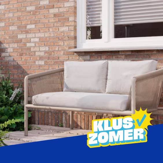 Beige tuinbank met zachte kussens voor een bakstenen muur en raam, omgeven door groen. Logo 'Kluszomer' rechtsonder.