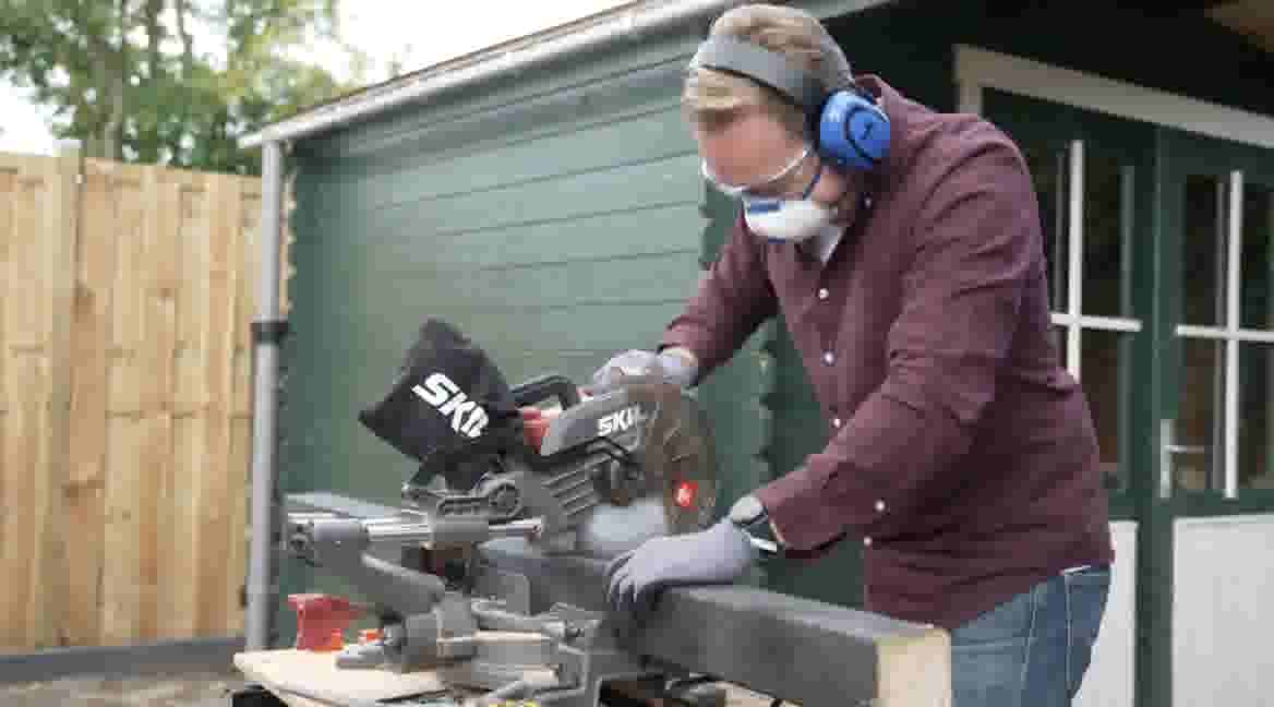 GAMMA klusser gebruikt een SKIL cirkelzaag met gehoorbescherming, veiligheidsbril en stofmasker bij groene schuur.