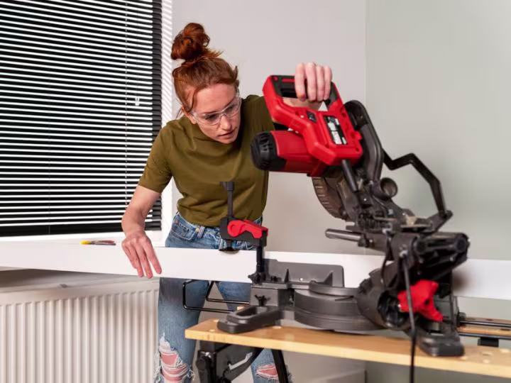 Persoon met veiligheidsbril gebruikt een rode elektrische zaag om hout te snijden op een werkbank bij een raam met jaloezieën.