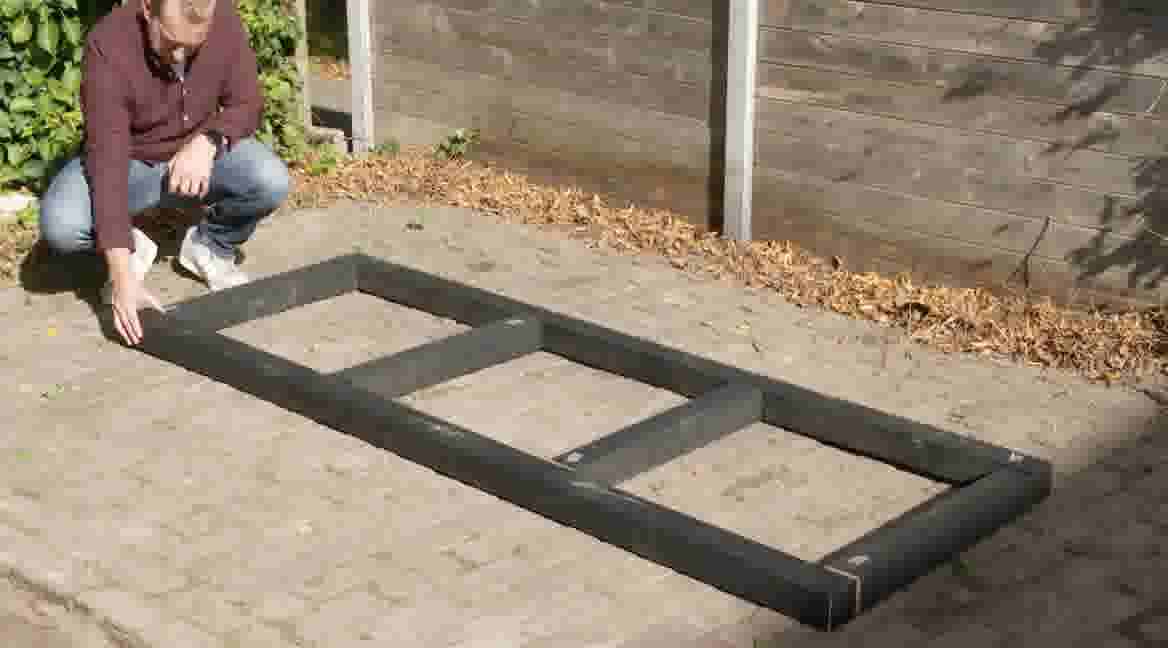 GAMMA klusser hurkt naast een rechthoekig zwart frame op bestrating in een tuin met houten schutting.