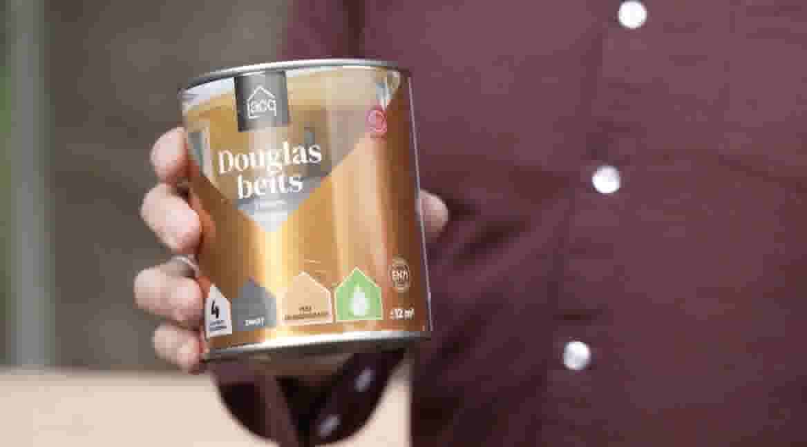 GAMMA klusser houdt een blik Douglas beits vast, met transparante afwerking en milieuvriendelijk label, tegen een bordeauxrode achtergrond.