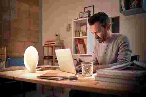 Man in grijze trui werkt geconcentreerd op laptop aan houten bureau met philips hue lamp en glas water in gezellige thuiswerkplek.