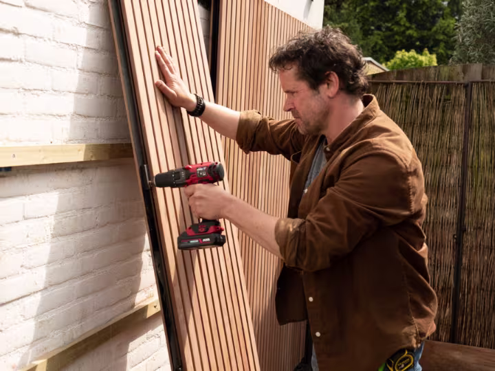Klusser bevestigt houten panelen aan witte muur met een accuboormachine in een tuin.