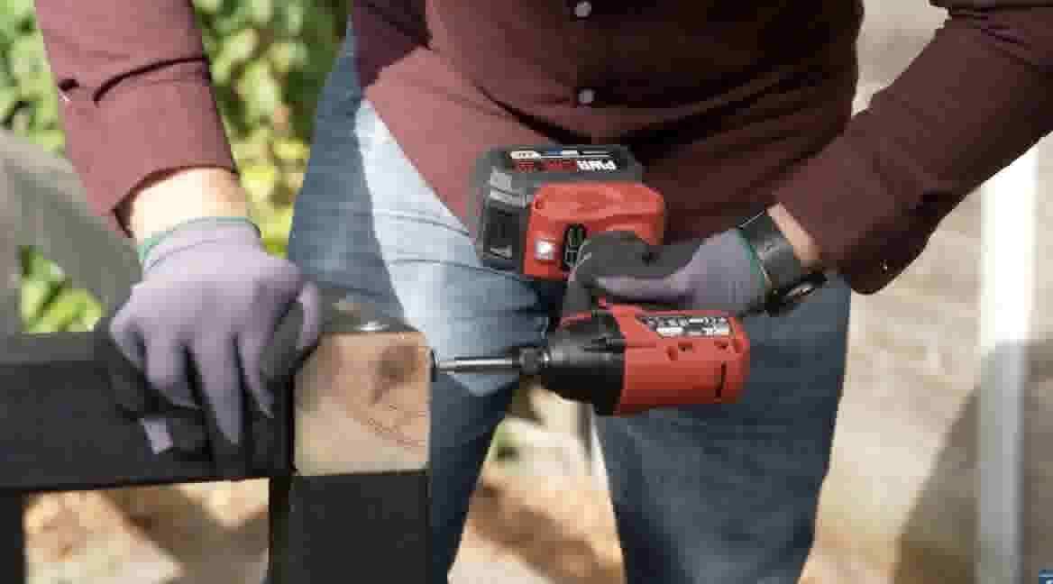 GAMMA klusser gebruikt een rode accuboormachine om een schroef in een houten balk te draaien, met werkhandschoenen aan.