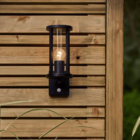 Zwarte buitenlamp met glazen cilinder en gloeilamp gemonteerd op houten schutting met bewegingssensor