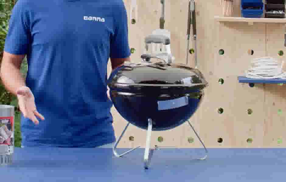 Persoon in blauw Gamma-shirt demonstreert een zwarte tafelmodel kogelbarbecue op een blauwe tafel, met houten wandpaneel op achtergrond.