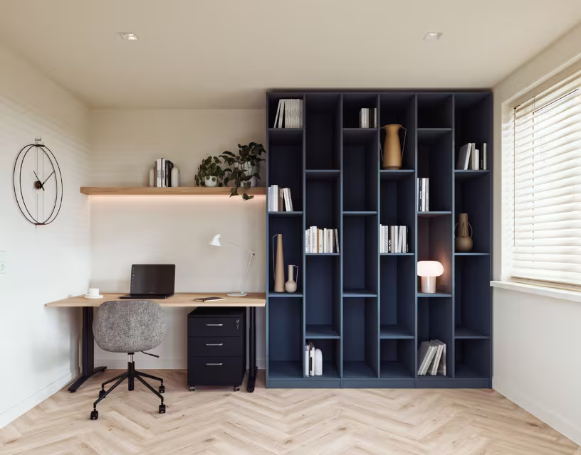 Modern thuiskantoor met houten bureau, grijze bureaustoel en donkerblauwe boekenkast met decoratieve items en boeken.