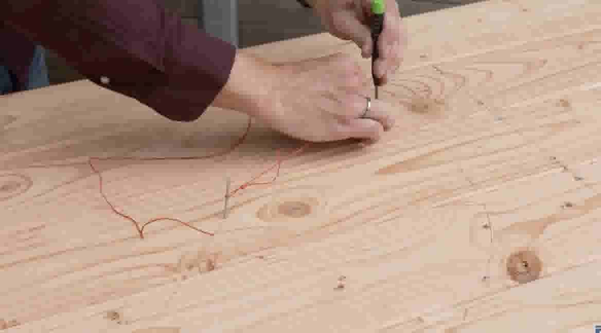 Handen markeren een houten oppervlak met een schroevendraaier, terwijl een rode draad over het hout loopt.
