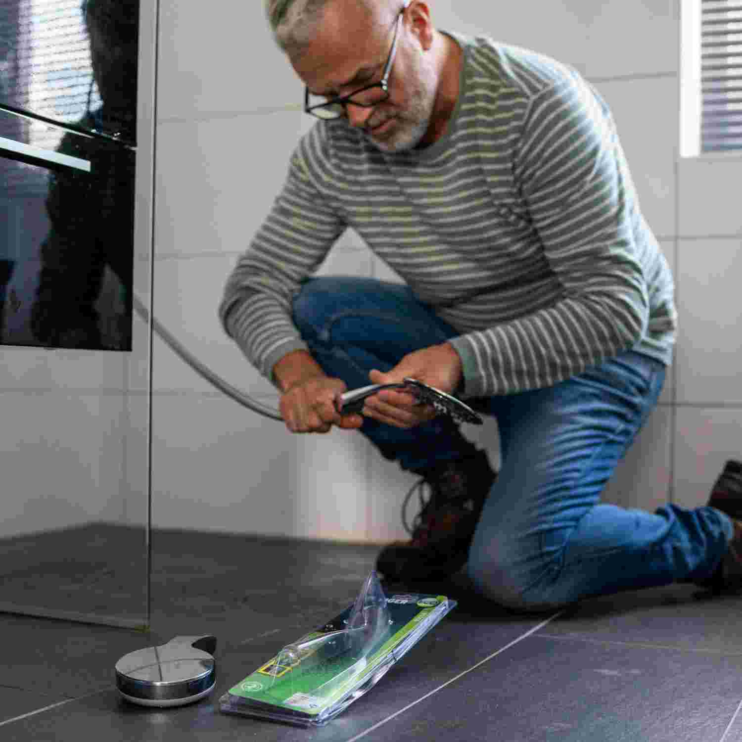 Man in gestreepte trui knielt op de vloer terwijl hij een waterbesparende douchekop installeert. 