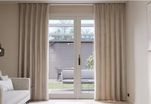 Woonkamer met beige gordijnen en glazen schuifdeuren die uitkijken op een terras met loungemeubels en tuin.