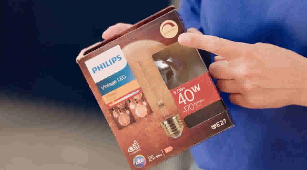 Persoon in blauwe GAMMA trui houdt een Philips LED lamp verpakking vast en wijst daarbij naar het icoon wat aangeeft dat de lamp dimbaar is. 