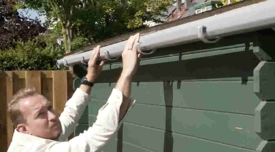 GAMMA klusser installeert een witte regenpijp op een groene schuur, met houten schutting en bomen op de achtergrond.