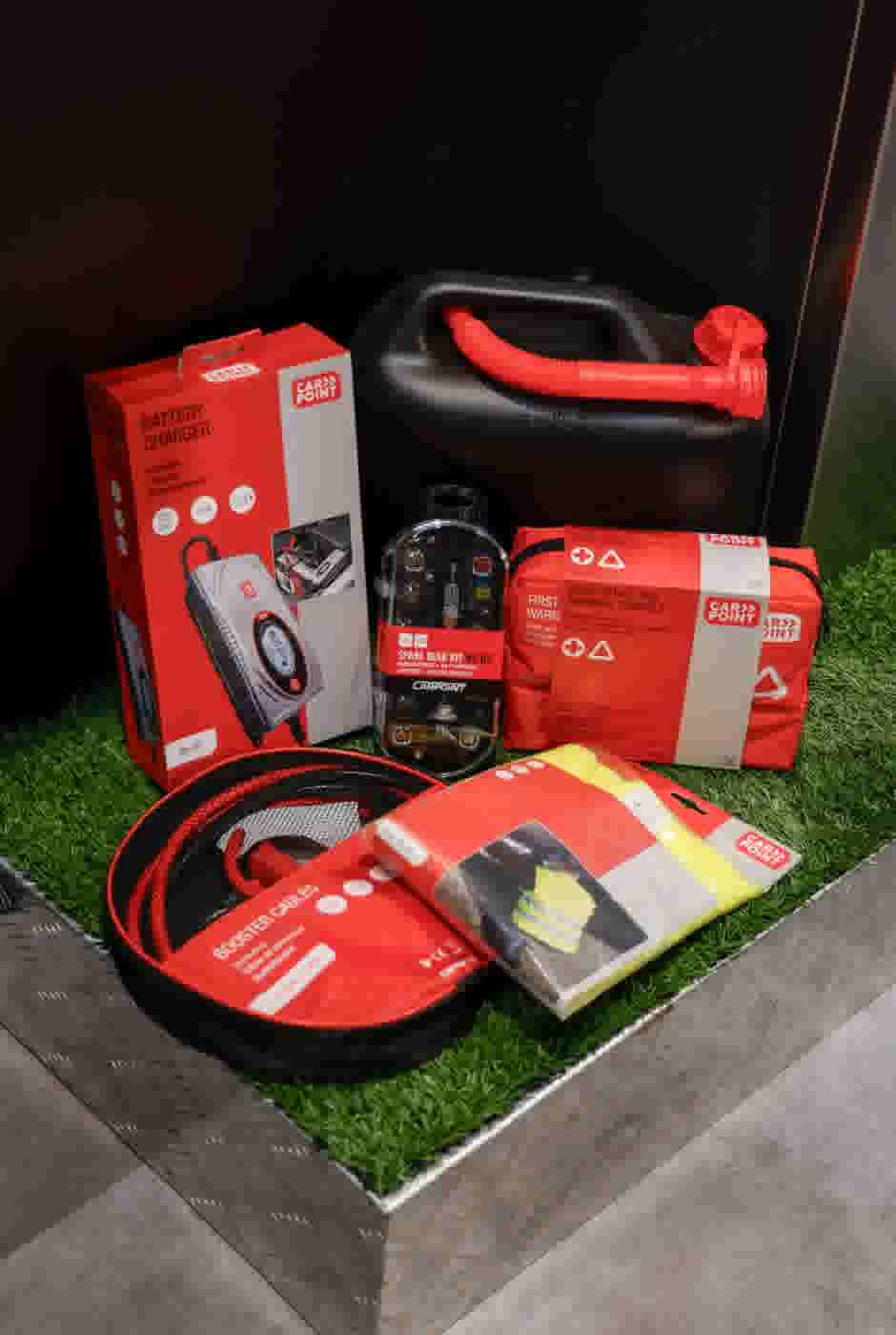 Auto-nooduitrusting met acculader, startkabels, jerrycan, reservelampenset en EHBO-kit op kunstgras geplaatst.