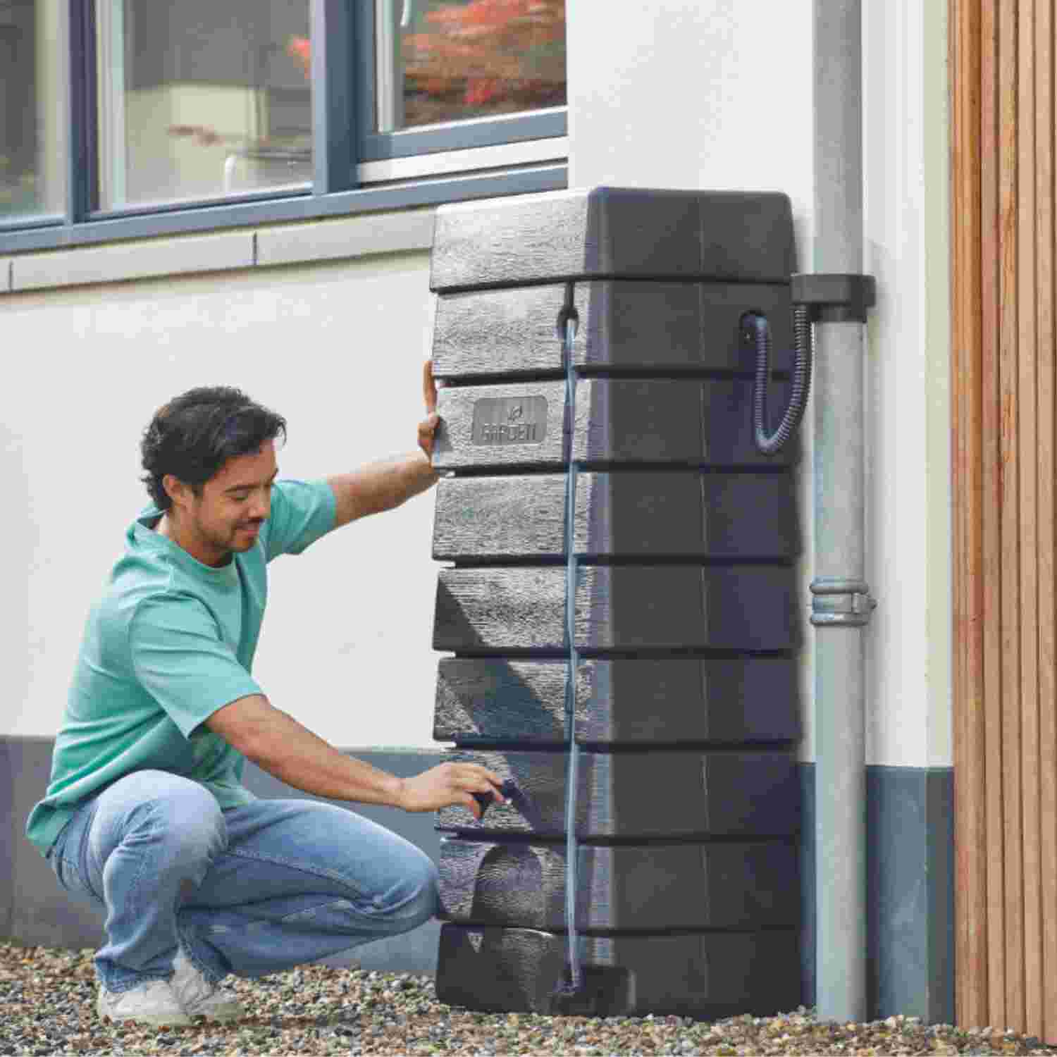 Man in turquoise shirt installeert een zwarte regenton tegen een witte muur naast een regenpijp.