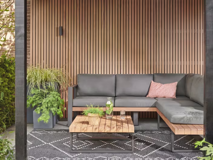 Moderne buitenlounge met grijze hoekbank, houten salontafel en verticale houten wandpanelen tegen een warme achtergrond.