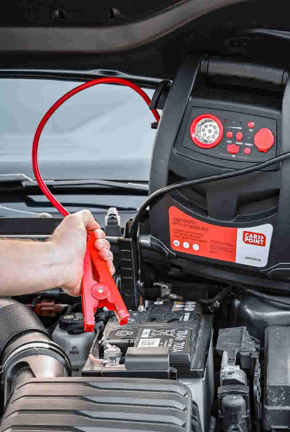 Hand bevestigt rode klem van een jumpstarter aan autoaccu, met CarPoint jumpstarter apparaat zichtbaar.