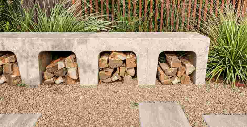Betonnen tuinbank met openingen voor brandhout, geplaatst op grind met siergrassen op de achtergrond.
