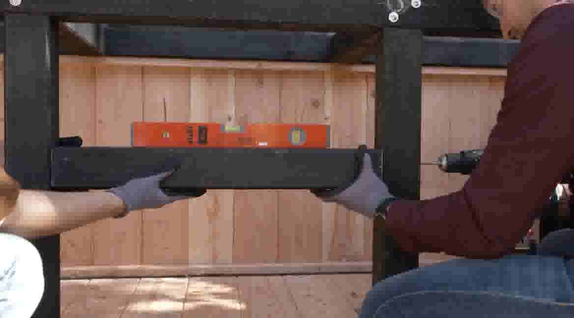 Twee personen in werkhandschoenen installeren een zwarte houten balk met een oranje waterpas en boormachine tegen een houten wand.