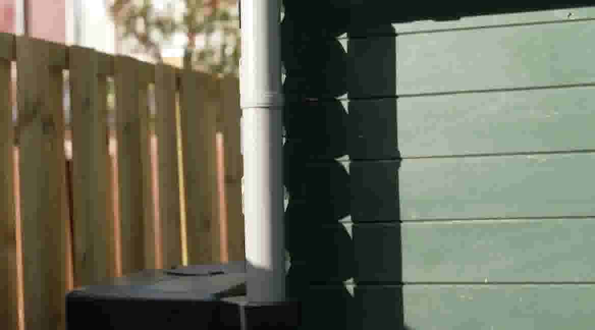 Houten schutting naast een groene muur met witte regenpijp, waarbij schaduwen op de muur vallen.
