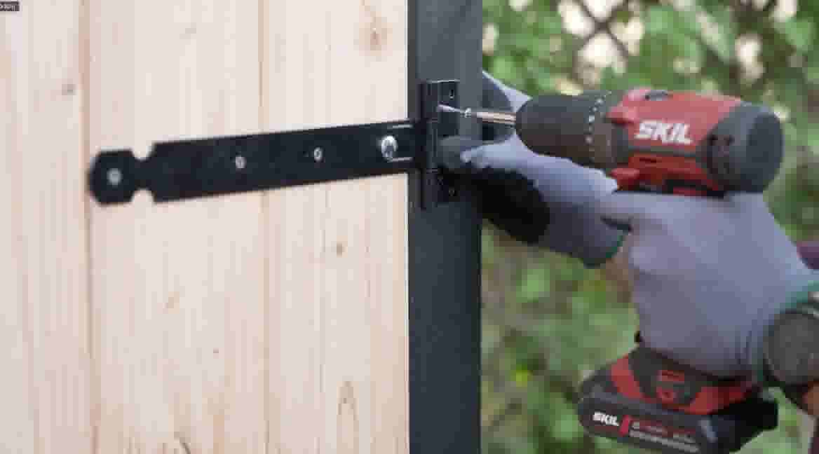 Gehandschoende handen gebruiken een rode SKIL boormachine om een zwart scharnier aan een houten schutting te bevestigen.