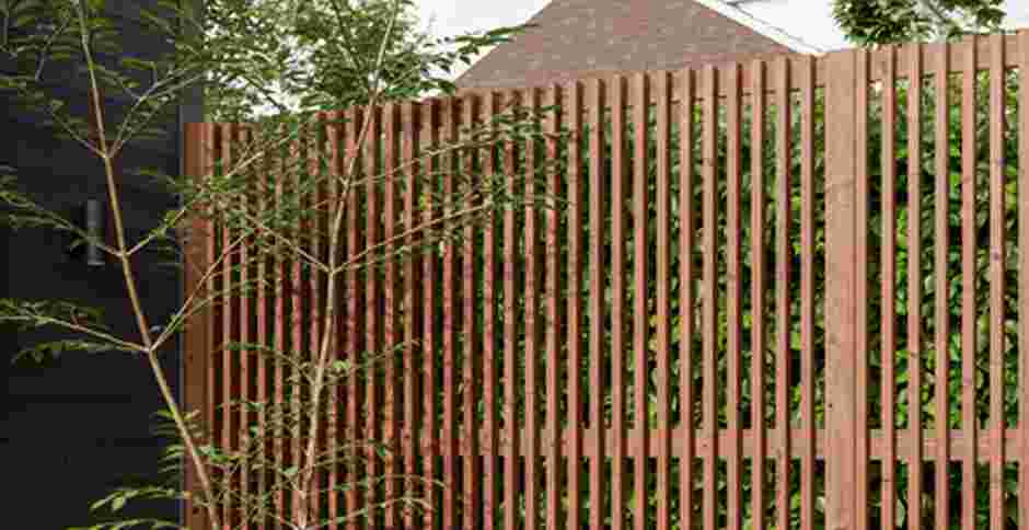 Moderne houten tuinafscheiding met verticale latten, groene planten groeien erachter, jonge boom links in beeld.