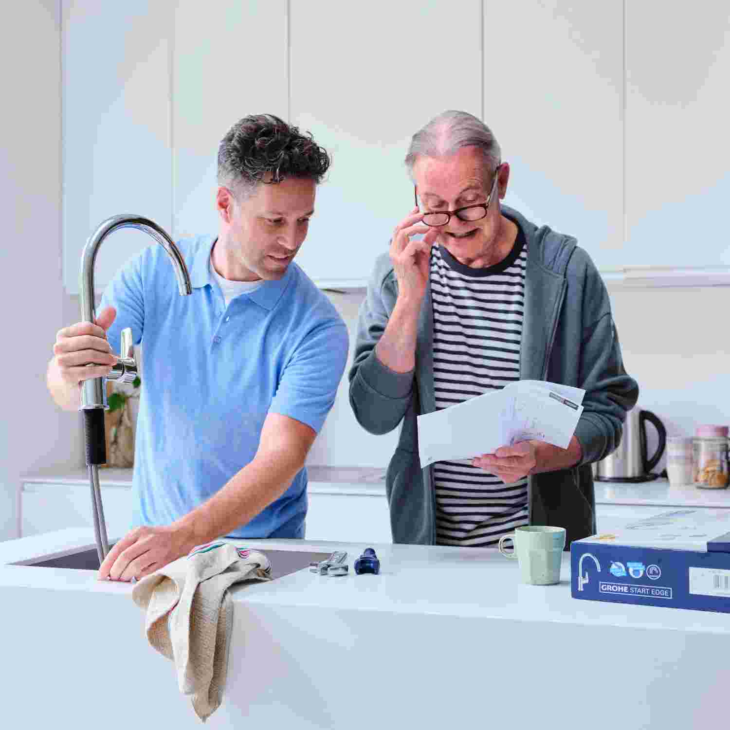 Jonge man in blauw shirt installeert een keukenkraan terwijl oudere man met bril instructies leest in een witte keuken.