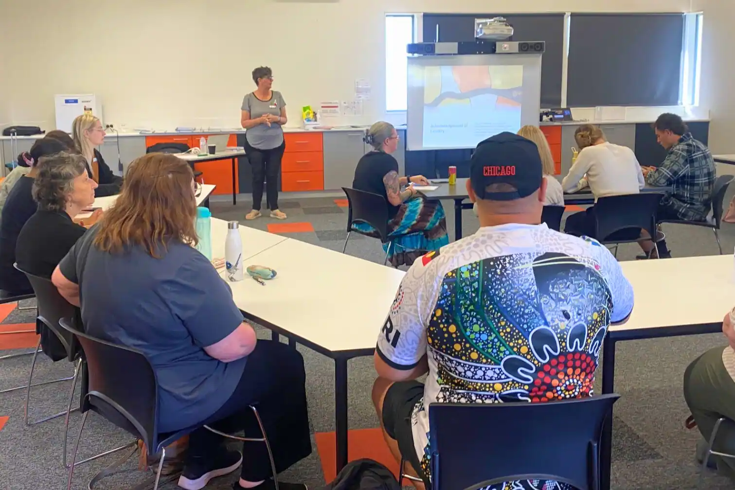 Group of adults seated around tables in a classroom, listening to Sue standing and presenting in front of a screen.