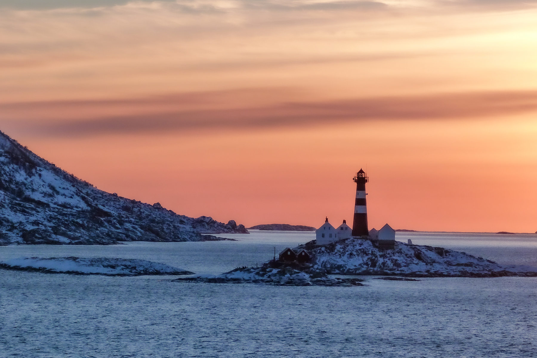 Bei Sonnenuntergang steht ein Leuchtturm auf einer verschneiten Insel, dessen Silhouette sich vor einem warm orange-rosa Himmel abzeichnet. Ruhiges Meer umgibt diese friedliche Szene.