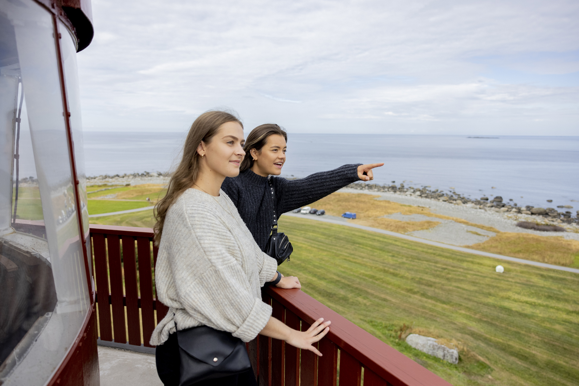 Two women stand on a lighthouse balcony, overlooking a serene coastal landscape. One points towards the sea, both appear relaxed and engaged.