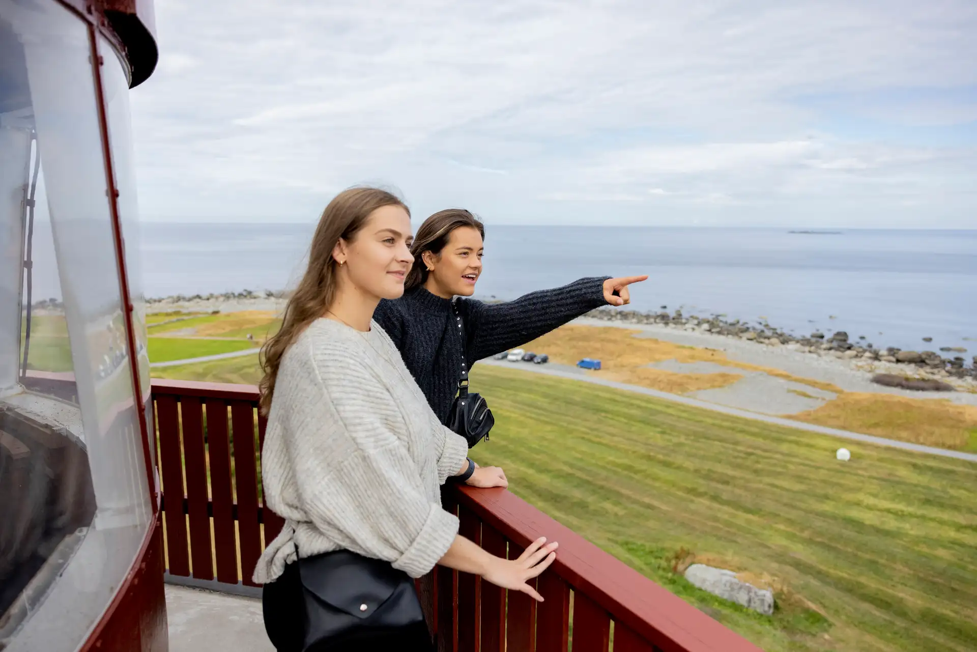 Zwei Frauen stehen auf dem Balkon eines Leuchtturms und blicken auf eine friedliche Küstenlandschaft. Eine zeigt auf das Meer, beide wirken entspannt und gebannt.