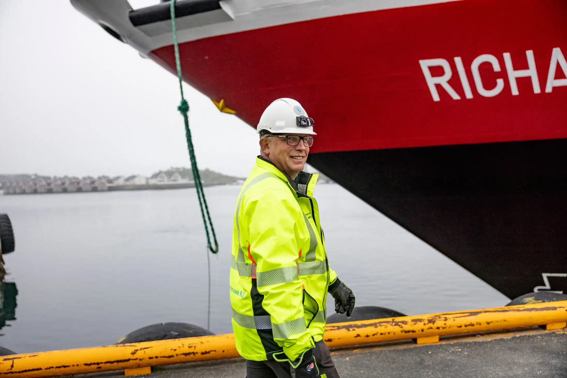 A man in a bright yellow safety jacket and white helmet smiles while standing beside a large red ship docked at a harbor on a foggy day.