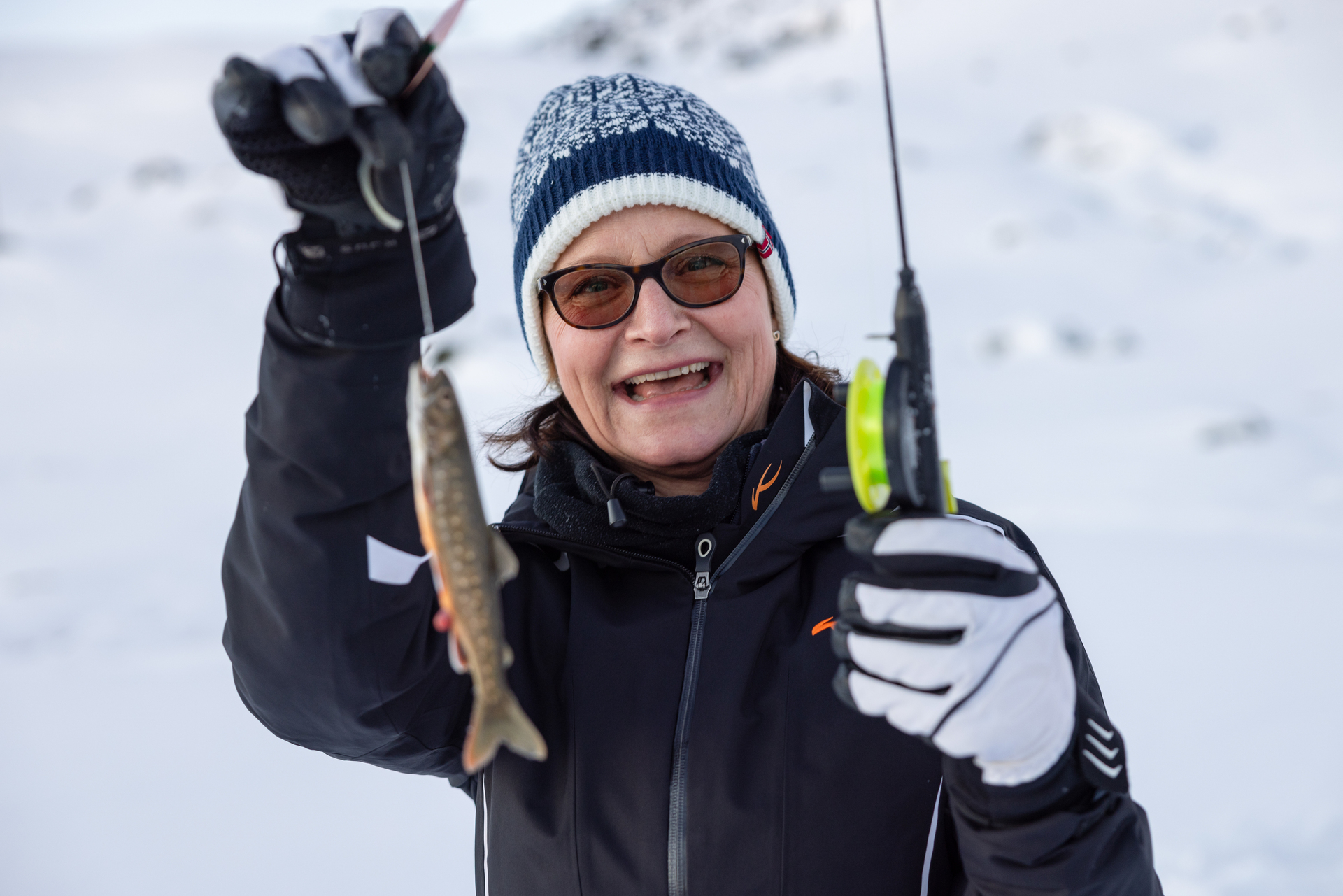A person wearing glasses and winter gear smiles while holding up a small fish and an ice fishing rod. The snowy landscape suggests a cold environment.