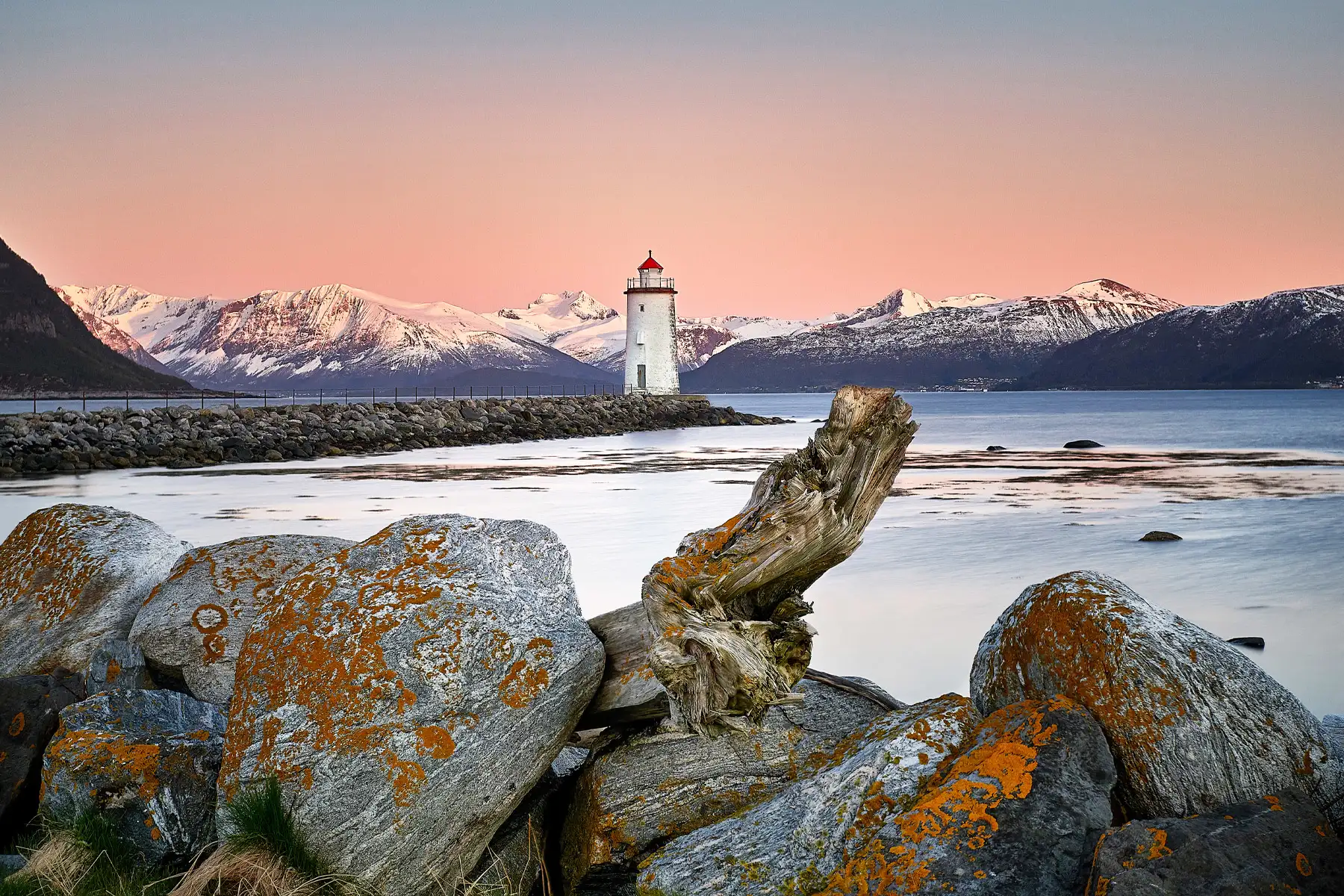 Ein weißer Leuchtturm mit rotem Dach steht an ruhigen Gewässern, eingerahmt von schneebedeckten Bergen und einem rosafarbenen Himmel bei Sonnenuntergang, mit Felsen im Vordergrund.