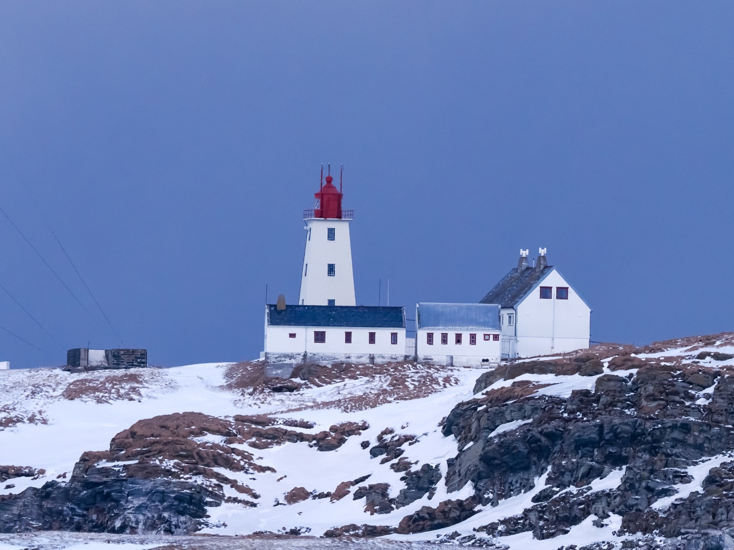 Ein weißer Leuchtturm mit rotem Dach steht auf einer schneebedeckten Klippe neben zwei miteinander verbundenen Gebäuden unter einem strahlend blauen Himmel und vermittelt ein Gefühl von Abgeschiedenheit und Ruhe.