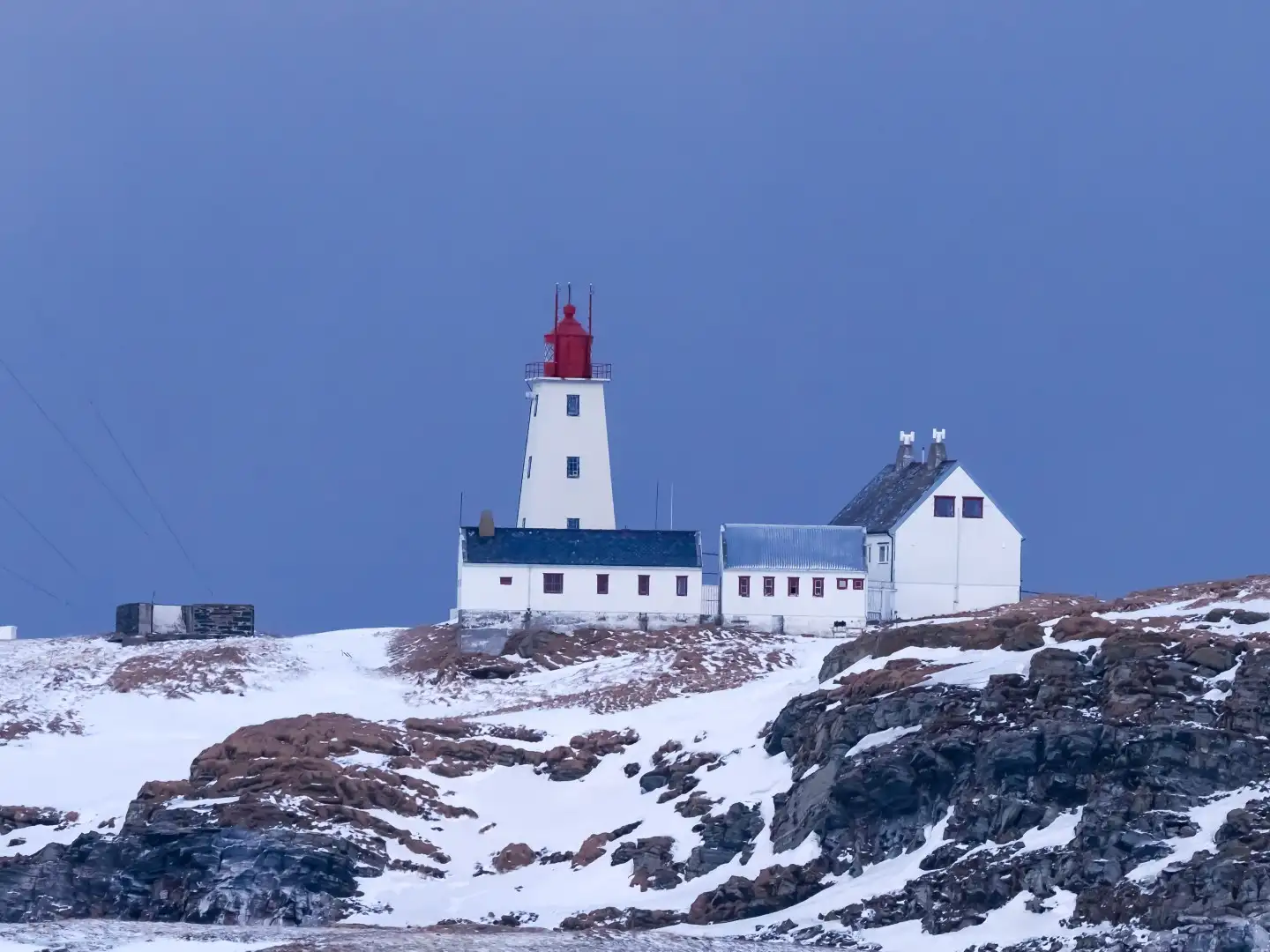 Ein weisser Leuchtturm mit rotem Dach steht auf einer schneebedeckten Klippe neben zwei miteinander verbundenen Gebäuden unter einem strahlend blauen Himmel und vermittelt ein Gefühl von Abgeschiedenheit und Ruhe.