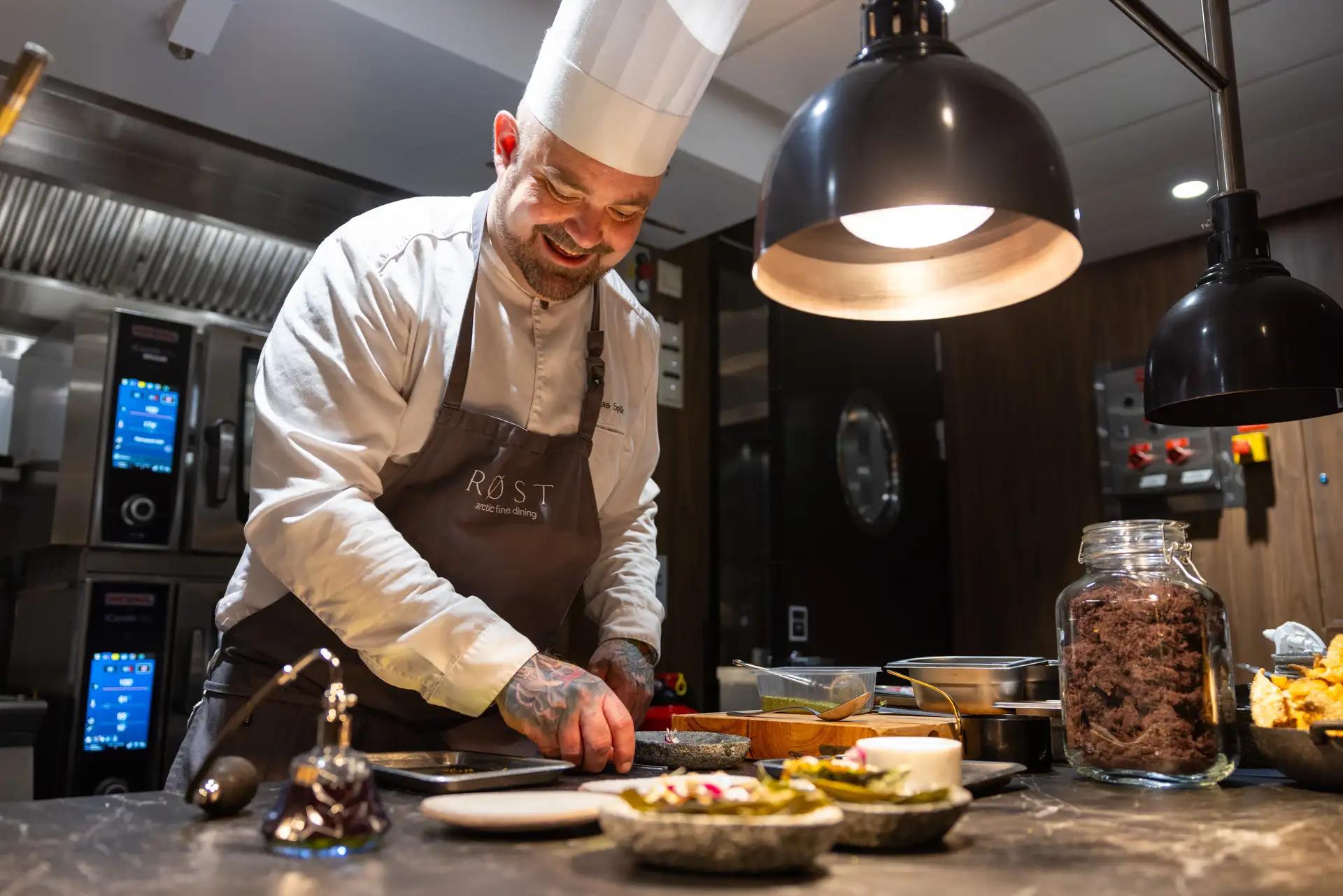 Chef in a white hat and apron joyfully plating food under warm lighting in a modern kitchen, surrounded by ingredients and cooking tools.