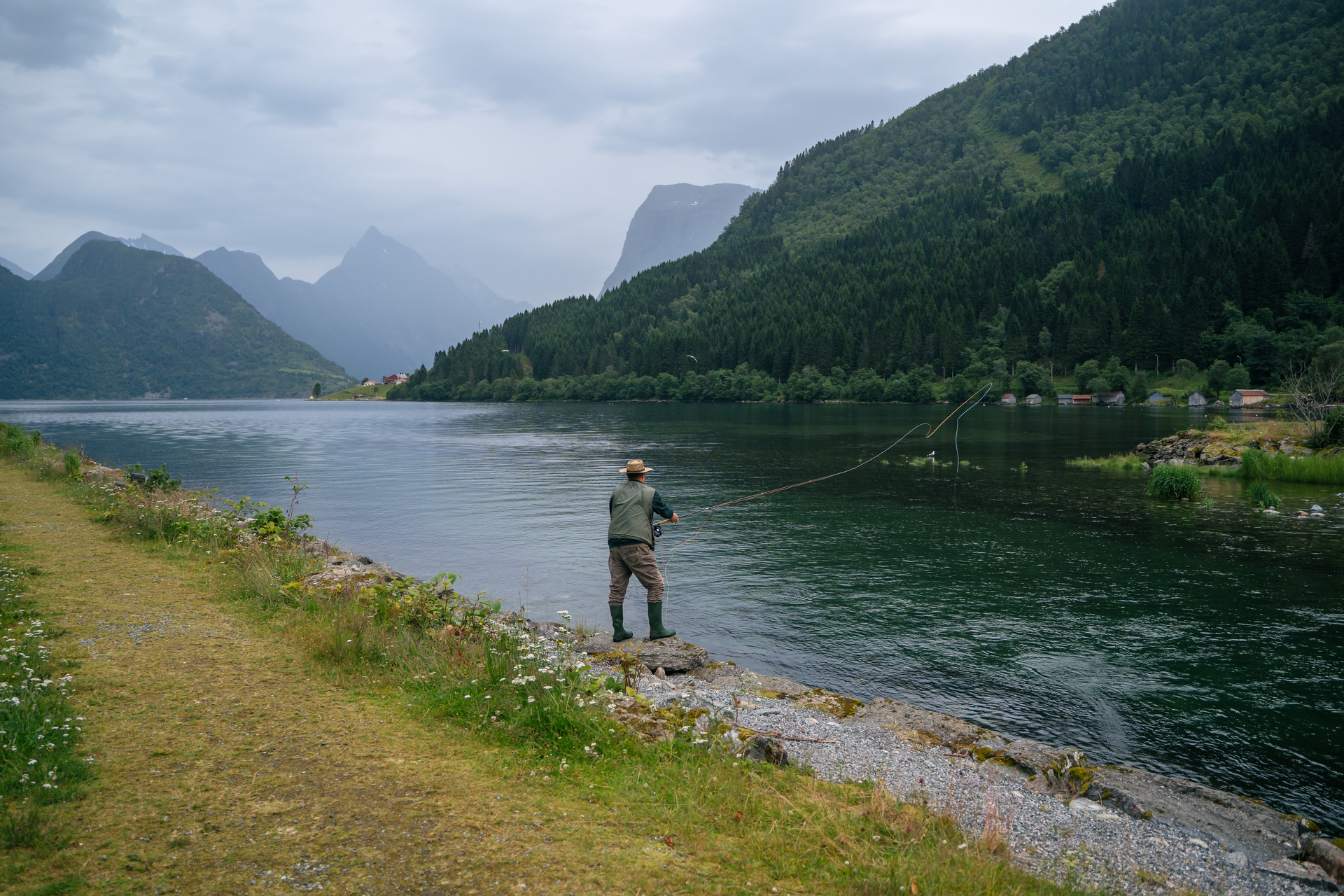A person in a hat fishes with a rod by a tranquil lake, surrounded by lush green mountains under an overcast sky, evoking a peaceful, serene mood.