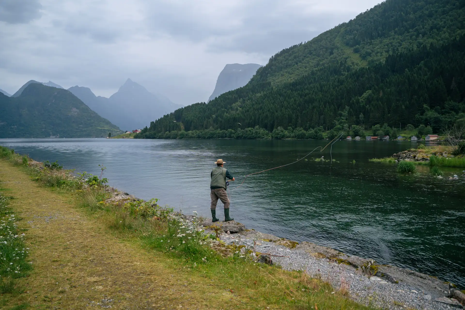 A person in a hat fishes with a rod by a tranquil lake, surrounded by lush green mountains under an overcast sky, evoking a peaceful, serene mood.