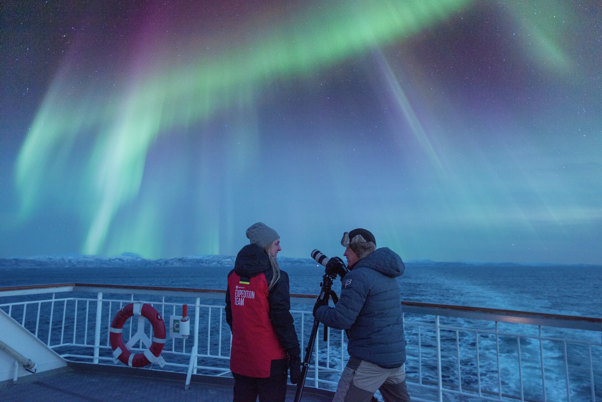 Guest photographing the Northern Lights with a tripod alongside a Hurtigruten expedition team member on deck of a Norway cruise ship