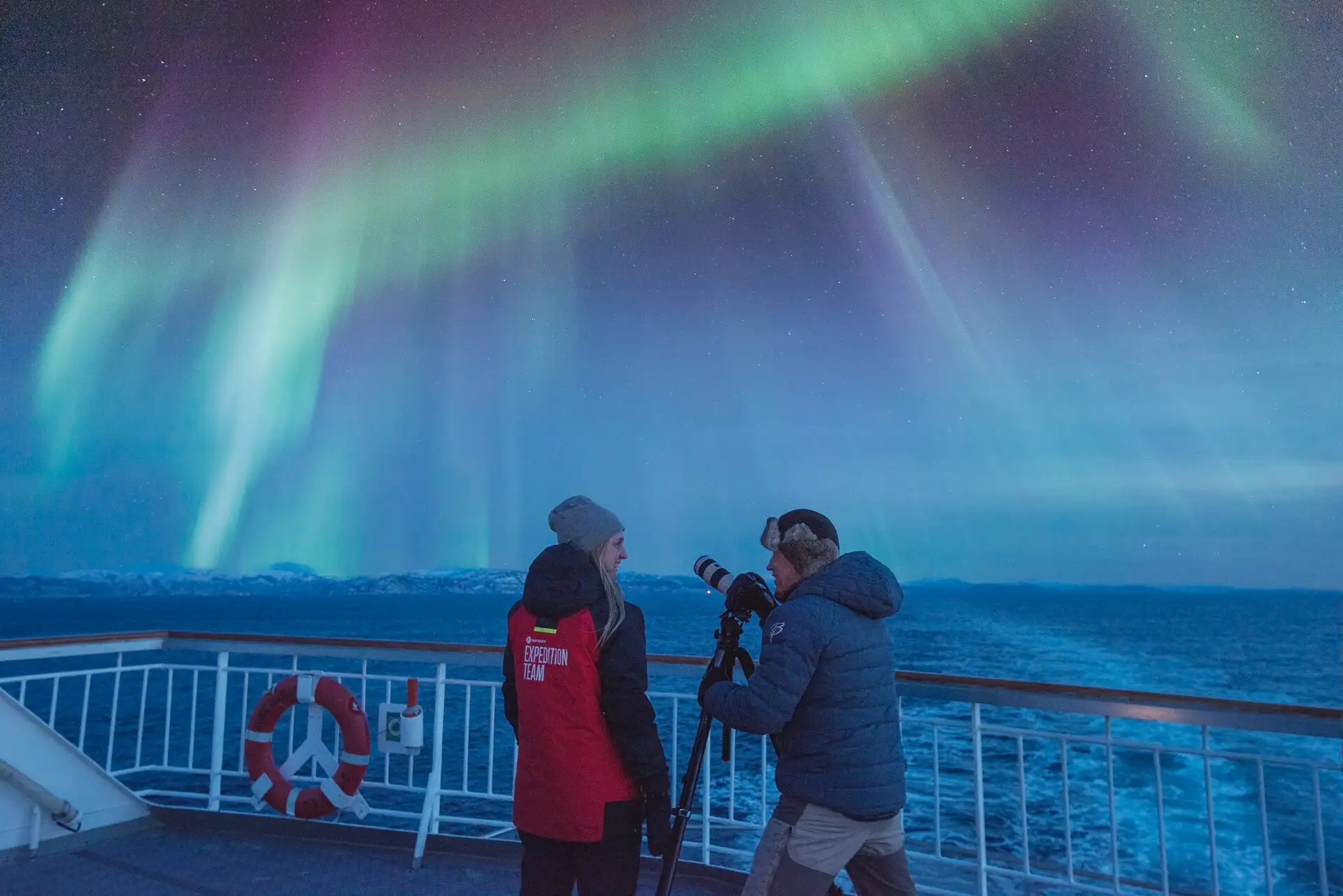 A tourist and crew member photographing the Northern Lights from the deck of a Hurtigruten ship